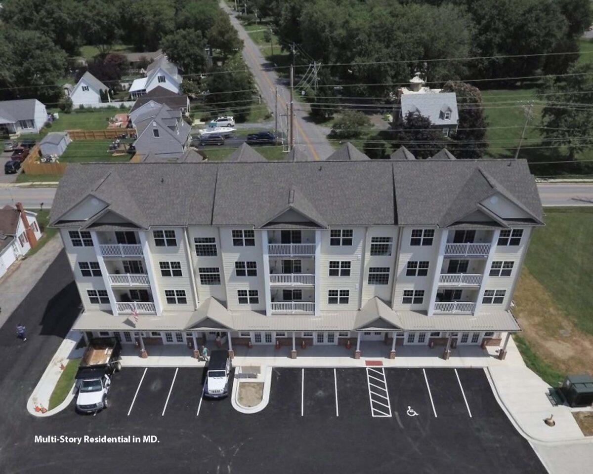 Aerial view of a multi-story residential building in Maryland with a gray roof and white exterior. The building has multiple balconies and an adjacent parking lot with several cars. Surrounding the building are other houses, trees, and a road.