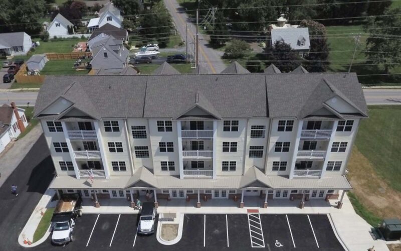 Aerial view of a multi-story residential building in Maryland with a gray roof and white exterior. The building has multiple balconies and an adjacent parking lot with several cars. Surrounding the building are other houses, trees, and a road.