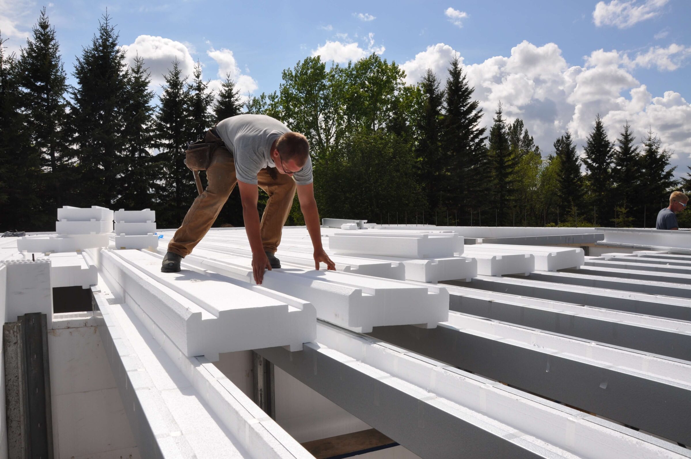 A construction worker bends over to position long white insulated panels on a building framework. The scene is outdoors, with dense, tall trees in the background under a partly cloudy sky.