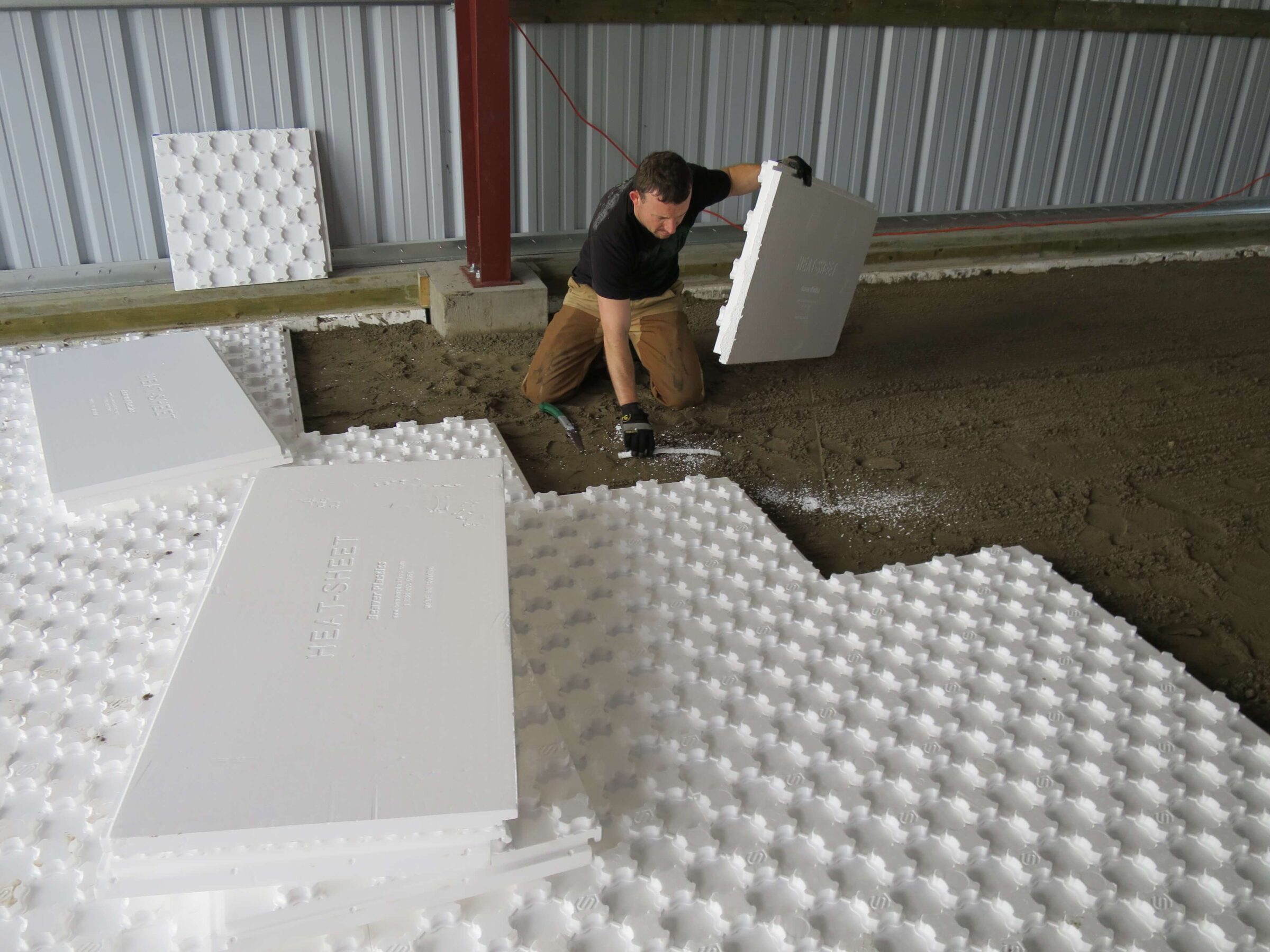 A construction worker is installing white EPS geofoam insulation panels on the ground inside a building. He is positioning a panel, aligning it with others. Additional panels are stacked nearby. The building's interior metal siding is visible in the background.