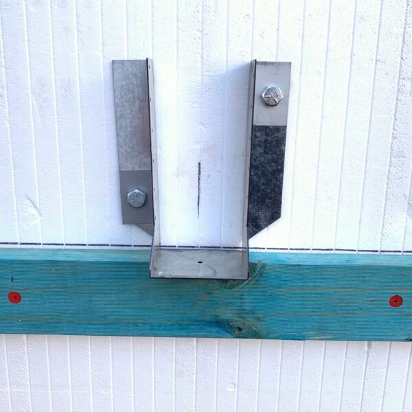 A metal bracket is mounted on a white corrugated surface, supported by a blue wooden plank with two visible red screws. The bracket consists of two vertical metal pieces attached together at the bottom, forming a U-shape.