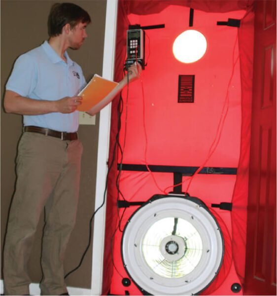 A man is conducting a blower door test inside a building. He holds a notepad and measuring device while standing beside a red door equipped with a large circular fan and a digital gauge. The setup is used to measure the air leakage in the building.