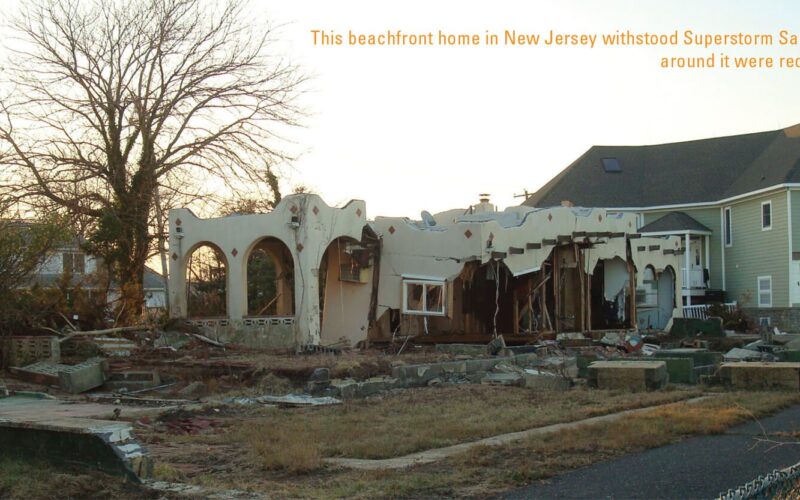 A lone standing beachfront home in New Jersey shows severe damage from Superstorm Sandy, partially destroyed with missing walls and debris scattered around. Surrounding houses and structures are reduced to rubble. Nearby, a larger, newer home appears intact.