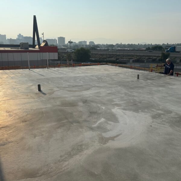 A large, freshly poured concrete slab on a rooftop with an industrial background. A person stands on the right side, observing. The city skyline is hazy, with several cranes visible in the distance under a clear sky.