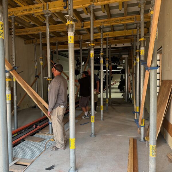 Construction workers standing among metal scaffolding beams inside a building under construction. The area is filled with wooden planks and other construction materials.