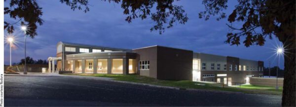 A modern, well-lit building at dusk with large windows and brick walls. The structure features a mix of flat and slightly angled roofs. Surrounding trees' branches frame the top of the image. An empty parking lot is visible in the foreground.