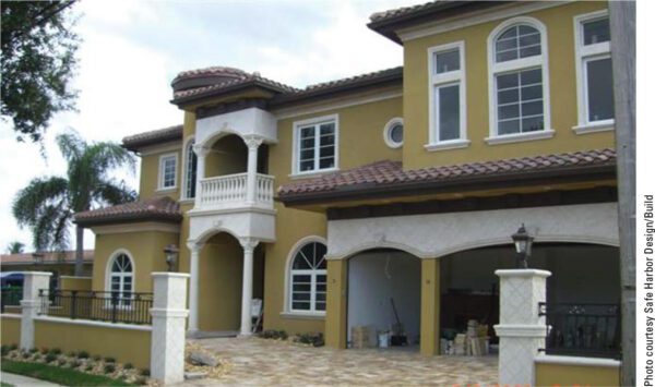 A two-story Mediterranean-style house with yellow stucco walls, white trim, arched windows, and a tiled roof. The front features a balcony with white columns, multiple garage doors, and a tiled driveway. Palm trees are visible on the left side.