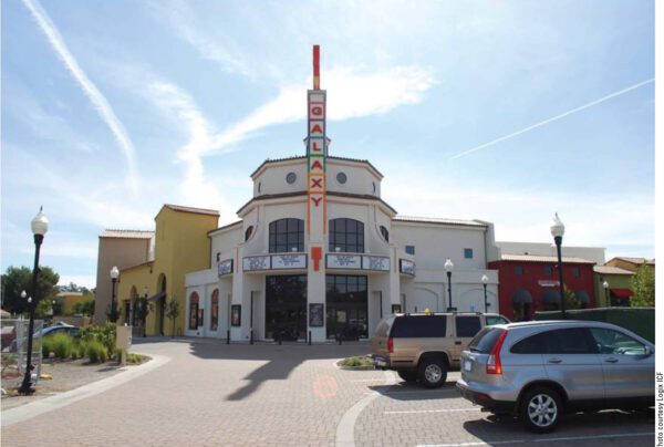 A modern cinema building labeled "Galaxy" stands under a clear sky. The structure has a prominent vertical sign, a large entrance, and is surrounded by light posts. Several cars are parked in the foreground on a cobblestone lot.