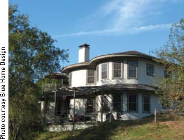 A two-story, modern-style house with large windows and a circular structure is surrounded by trees. The house is predominantly white, and a shaded patio area with outdoor furniture is visible on the ground floor. The sky above is clear and blue.