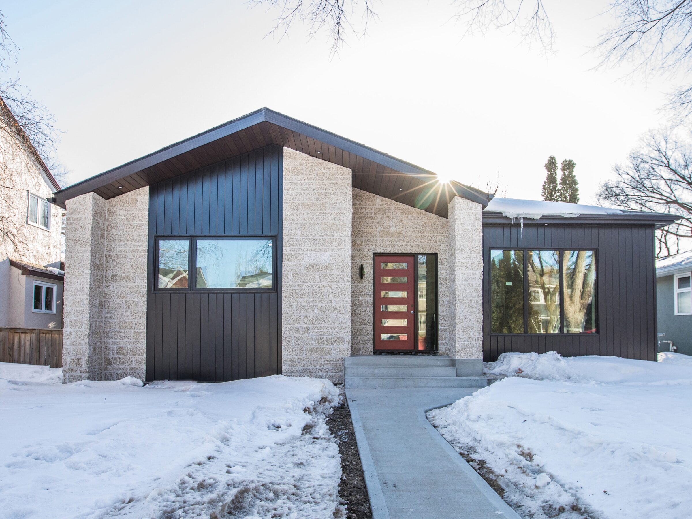 A modern house with contrasting dark brown and light stone exterior stands amidst a snowy landscape. The home features large windows, a sleek red door with glass inserts, and a concrete pathway leading to the entrance. Leafless trees are visible in the background.