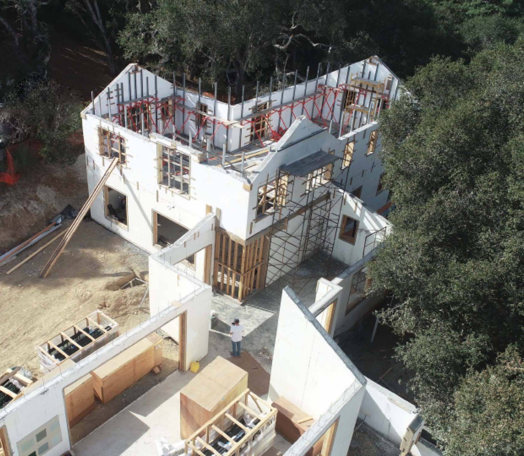 Aerial view of a large house under construction surrounded by trees. The walls and framework are partially completed, and scaffolding is visible on the structure. Construction materials and tools are scattered around the site, with one worker standing near the building.